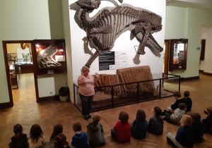 Na ścianie znajduje się odcisk szkieletu dinozaura. Po prawej stronie stoi kobieta - pracownik muzeum. Przed nią siedzą w szeregu dzieci. Dzieci są odwrócone tyłem i patrzą na kobietę. Po lewej i po prawej stronie widać inne eksponaty muzealne.