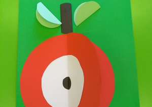 Półprzestrzenne jabłko w kolorze czaerwonym z białym środkiem w kształcie kółka z czarną kropką pośrodku, brązowym ogonkiem i dwoma zielonymi listkami. Jabłko przyklejone jest do zielonej kartki papieru.