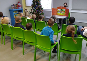 Na zielonych krzesłach siedzi 12 dzieci tyłem do obiektywu, patrzą na drewniany teatrzyk, wewnątrz którego umieszczona jest ilustracja. Za teatrzykiem siedzi kobieta. Po lewej stronie widać dużą choinkę ozdobioną bombkami i niski regał z książkami.