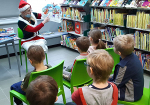 Na zielonych krzesłach siedzi 6 dzieci, tyłem do obiektywu. Patrzą na obrazek w książce, którą trzyma kobieta przebrana za elfa. Wskazuje ona palcem dużego bałwana. Po prawej stronie widać wysokie regały z książkami.