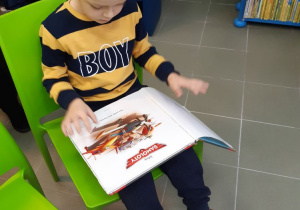 Na zielonym krześle siedzi chłopiec. Na kolanach trzyma otwartą książkę. Na perwszej stronie widać napis "samoloty" i obrazek z samolotami. Chłopiec ubrany jest w bluzę w czarno-żółte pasy z napisem "boy"
