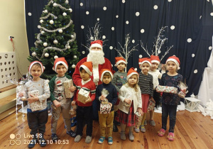 W centralnej części zdjęcia siedzi Mikołaj, przed nim stoi dziewięcioro dzieci z oddziału II. Trzymają prezenty. Po lewej stronie stoi choinka i renifer, w tle granatowa kurtyna z białymi kulkami imitującymi śnieg.