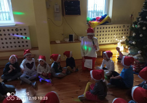 Grupa dzieci siedzi w kole na podłodze, każdy ma na głowie czerwoną czapkę z białym obszyciem i pomponem. Pomiędzy dziećmi siedzi pani Dyrektor, w jednym ręku trzyma mikrofon przez który mówi, a w drugim kartkę papieru z tekstem. W tle widać fragment choinki, okna i sprzęt muzyczny. Jest półmrok.