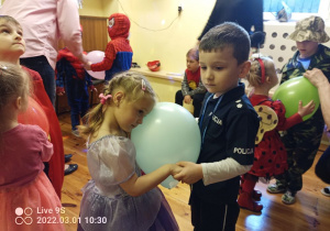 Na zdjęciu widać tańczące dzieci przebrani za różne postacie. W centralnej części zdjęcia tańczy chłopiec z dziewczynką, trzymając swoimi brzuchami duży balon. Dziewczynka jest w stroju księżniczki a chłopiec policjanta.