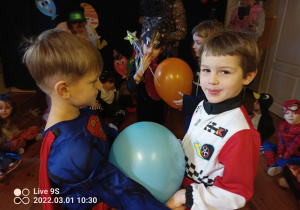 Dwóch chłopców tańczy trzymając się za ręce, między nimi jest balon. Jeden z chłopców przebrany jest za supermena a drugi za kierowcę formuły 1.
