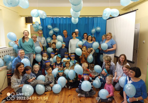 Grupa osób dzieci i dorosłych ubrana na niebieski na tle niebieskiej kotary i niebieskich balonów zwisających z sufitu. Wszyscy trzymają niebieskie balony.