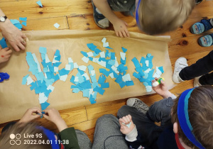 Na szarym kartonie kontury czterech dłoni wyklejone skrawkami niebieskiego papieru. Dookoła siedzą dzieci, mają pochylone głowy nad obrazkiem.