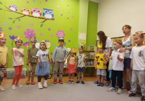 W sali na tle zielonej ściany z kolorowymi naklejkami sów stoi w półkolu grupa dzieci. Są w ruchu. Na głowach mają opaski z wizerunkami zwierząt. Za nimi stoją dwie dorosłe kobiety.