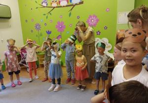 Grupa dzieci z opaskami z wizerunkami zwierząt na głowie stoi w półkolu. Część z nich trzyma dłonie zwinięte w rulon przy oczach. Za nimi stoi dorosła kobieta także trzymając dłonie przy oczach.
