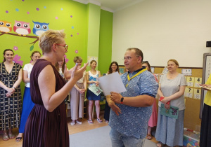 Pani Dyrektor stoi przez mężczyzną, który trzyma w rękach kartkę papieru. Na jego szyi wisi medal na żółtej wstążce.