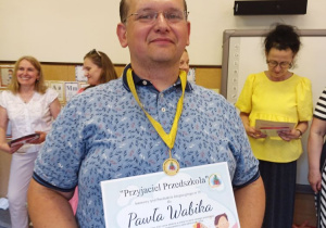 Na środku zdjęcia stoi mężczyzna trzymając na wysokości brzucha dyplom z napisek Przyjaciel przedszkola. Na szyi na zawieszony medal na żółtej wstążce. Uśmiecha się.