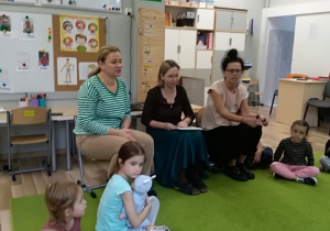 Na pierwszym planie widać trzy nauczycielki, które siedzą na krzesełkach. Po prawej i po lewej stronie na dywanie siedzą dzieci.