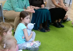Trzy nauczycielki siedzą na krzesełkach. Przed nimi na dywanie siedzi dziewczynka, która trzyma lalkę. Po lewej stronie widać fragmenty postaci siedzących dzieci.