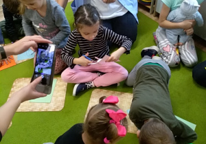 Na zdjęciu widać grupę dzieci, które siedzą na dywanie. Jedna z dziewczynek trzyma kartkę i nożyczki. Chłopiec i dziewczynka pochylają się na koszykiem z nożyczkami. W tle widać osobę dorosła i dziewczynkę trzymającą lalkę.