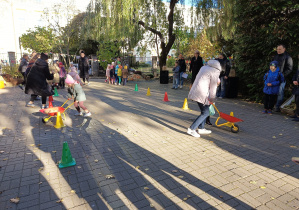 Zdjęcie przedstawia dzieci podzielone na dwie drużyny. Przed nimi stoją rozstawione kolorowe pachołki. Jedno dziecko z każdej drużyny pcha pomiędzy pachołkami czerwoną taczkę. Taczka jest wypełniona ziemniakami.