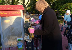 Zdjęcie przedstawia czerwoną maszynę do popcornu. Obok niej stoi kobieta nakładająca łopatką popcorn do pojemnika. Za nią stoją dzieci ustawione w kolejkę.